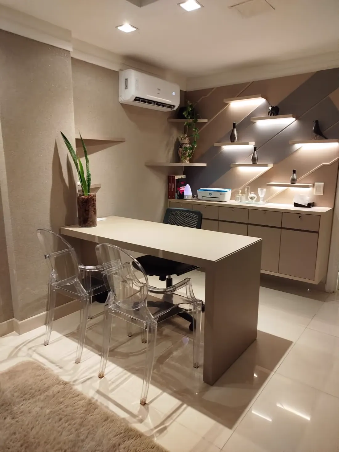Consultório com mesa clara, cadeiras transparentes e prateleiras iluminadas